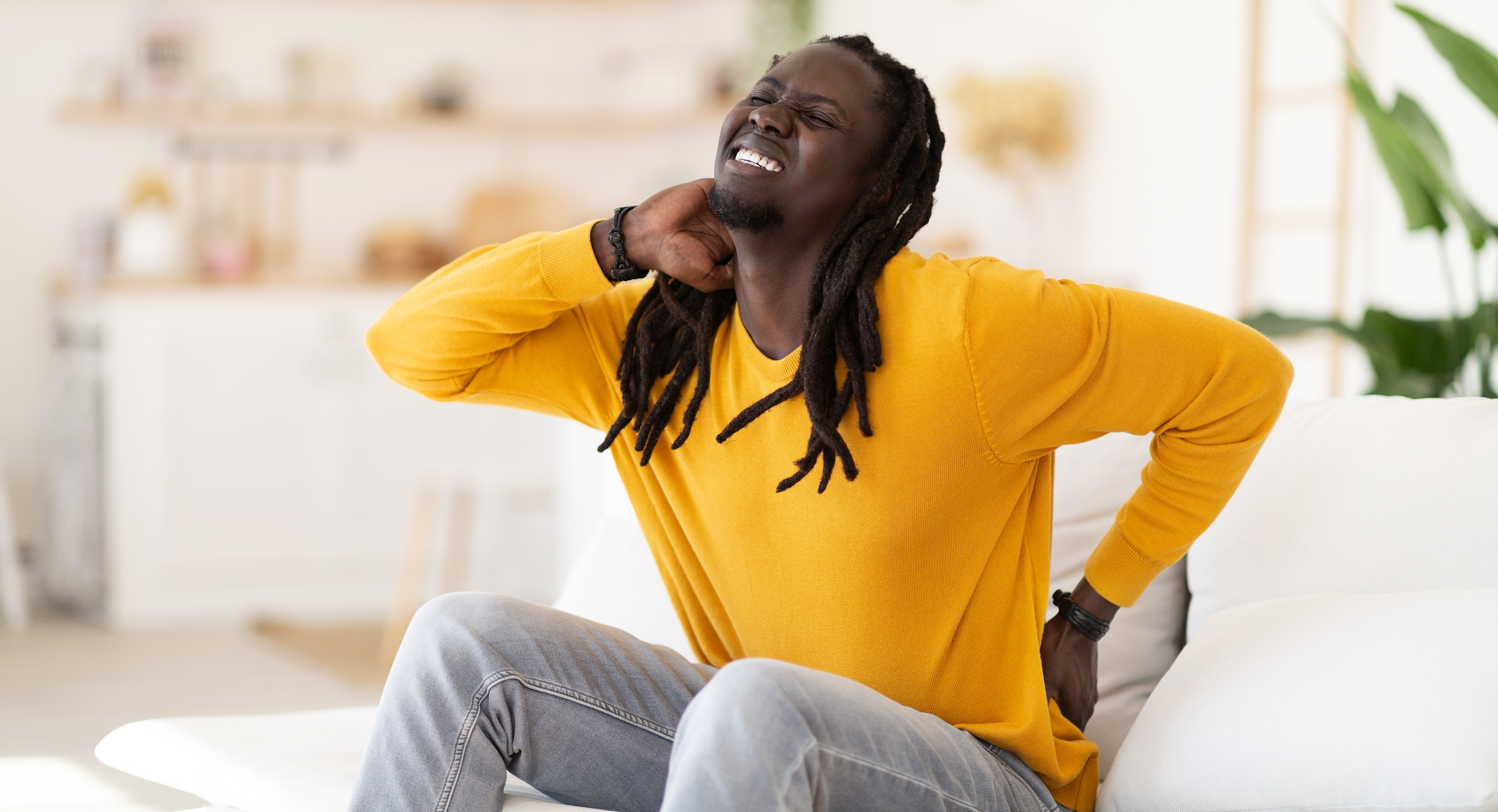 Image of a man sitting on a sofa holding his back and neck as if in pain